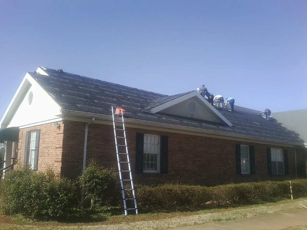 Crew tearing shingles off a brick ranch roof with a ladder set — Emergency Roof Repair work by our crew in Pearsall
