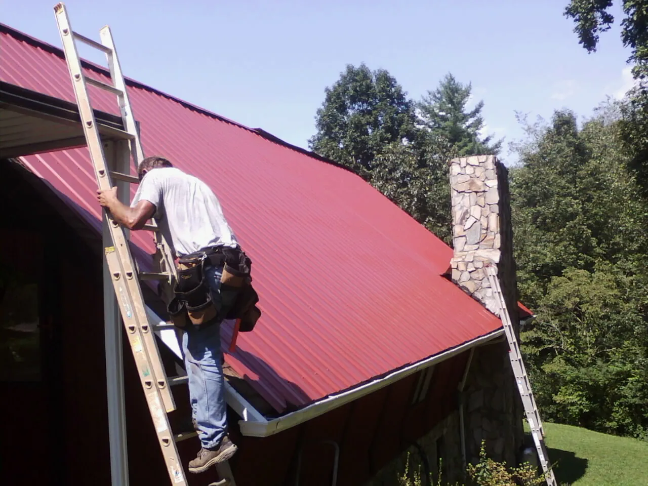 Roofer on a ladder beside a red metal roof and stone chimney — Soffit & Fascia Repair by Roofing Brothers in Pearsall